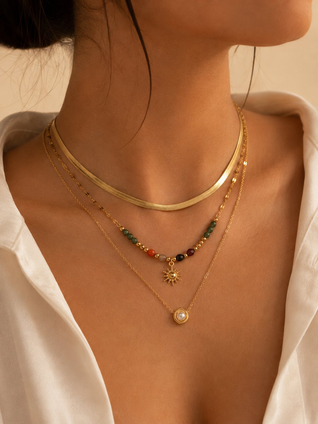A close-up of a model's neck and chest wearing a layered gold necklace set. The set includes a thick gold snake chain, a delicate gold chain with a small gold sun pendant and a pearl bead, and a gold chain with small red, green, and black beads.