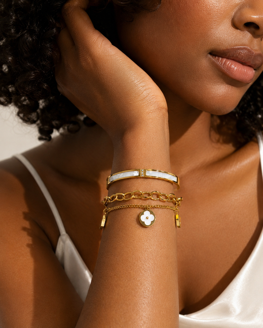 A close-up of a woman wearing a stack of gold bracelets on her wrist, including a bangle with a white inlay and a small white clover charm, against a warm neutral background.