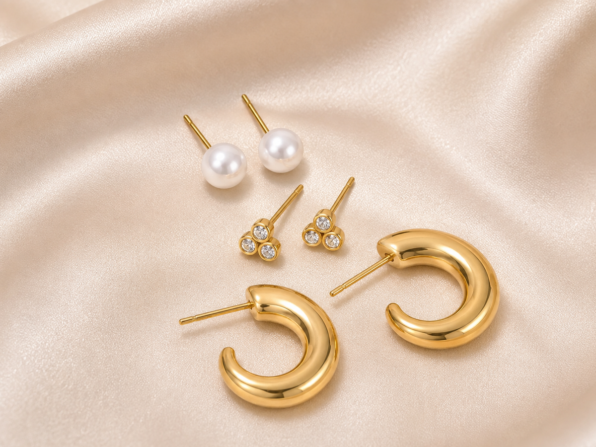 A set of gold and pearl stud earrings arranged on draped champagne satin fabric. The set includes a pair of classic white pearl studs, a pair of small gold tri-oval stud earrings with clear stones, and a pair of chunky open C-shaped gold hoop earrings.