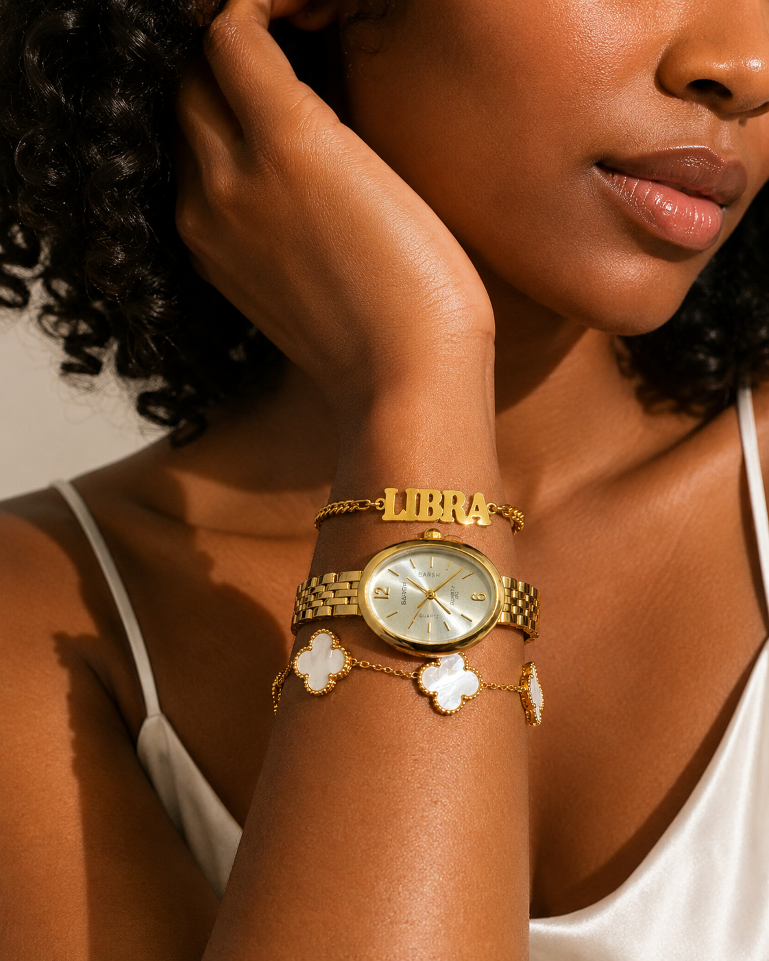 A close-up lifestyle shot of a woman wearing a gold-tone watch and matching bracelets on her wrist. The watch has a gold-tone metal link band, a round off-white dial, and a gold-tone bezel.