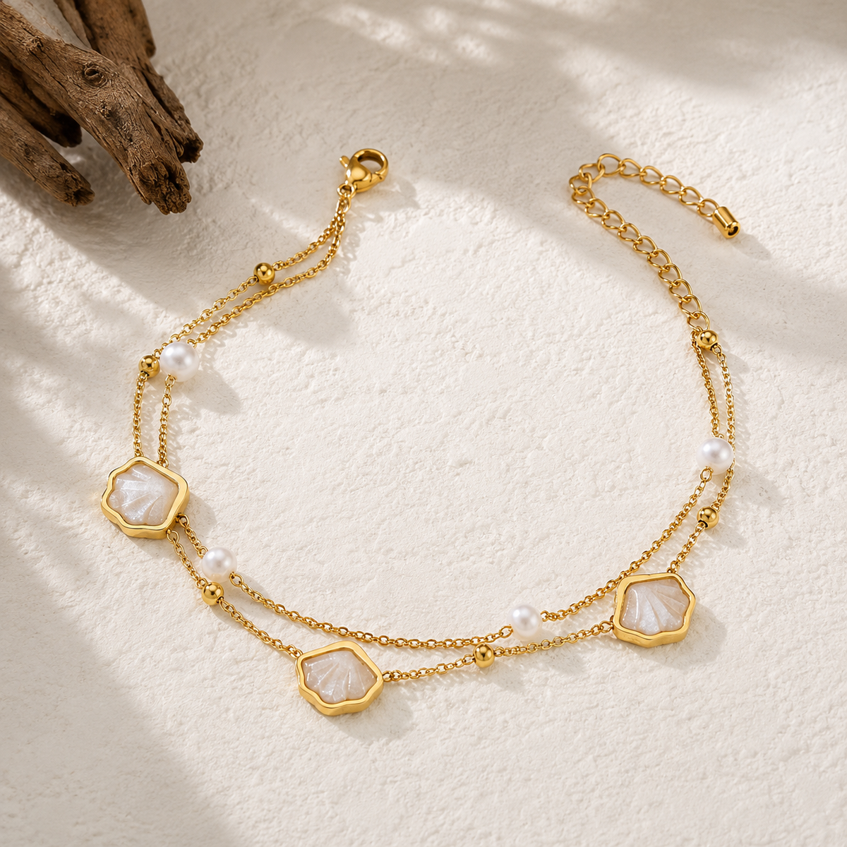 A gold-tone double-strand anklet featuring three octagonal mother-of-pearl bead charms and small white pearl beads, displayed on a textured white surface with a piece of driftwood in the corner.