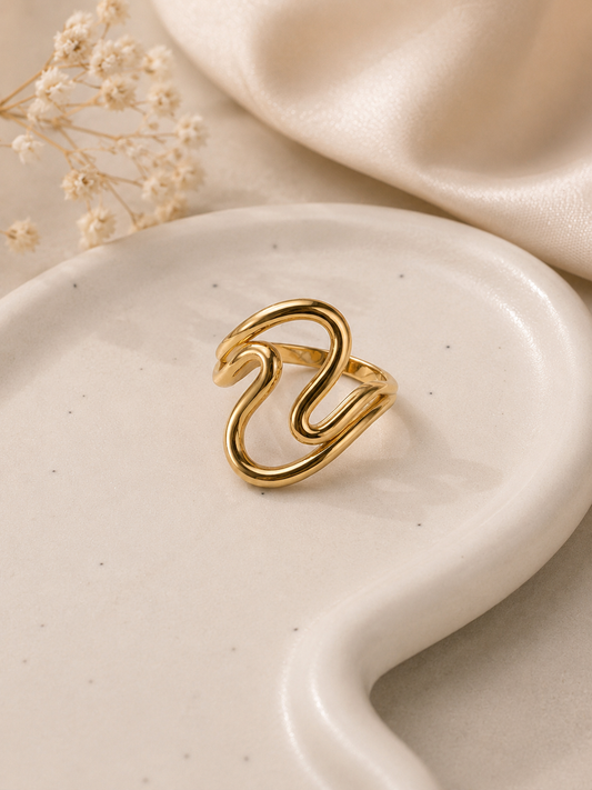 A gold-plated swan geometric ring displayed on a white ceramic dish with a beige satin fabric and dried baby's breath flowers in the background.