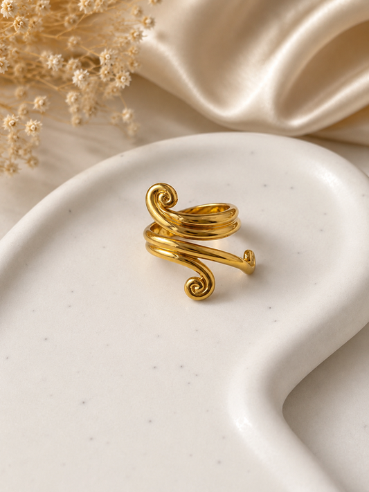 A gold-toned spiral wrap ring displayed on a white speckled ceramic dish, with soft beige satin fabric and dried floral stems in the background.