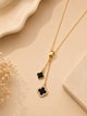 A gold-tone lariat necklace with two clover-shaped pendants, one solid black and one black with a border of small clear stones, displayed on a beige surface with dried flowers.