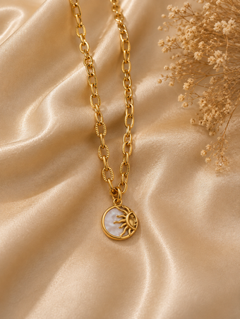 A gold chain necklace with a circular pendant featuring a gold sun design over a white mother-of-pearl background, styled on draped champagne satin fabric with dried flowers.