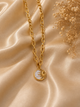 A gold chain necklace with a circular pendant featuring a gold sun design over a white mother-of-pearl background, styled on draped champagne satin fabric with dried flowers.
