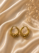 A pair of gold hoop earrings with a textured, beaded lower half, displayed on draped champagne-colored satin fabric with dried baby's breath flowers.