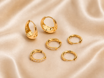 Six gold huggie hoop earrings, including two with small clear pavé accents and two with a single round clear stone, arranged on a draped beige satin fabric background.