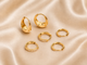 Six gold huggie hoop earrings, including two with small clear pavé accents and two with a single round clear stone, arranged on a draped beige satin fabric background.
