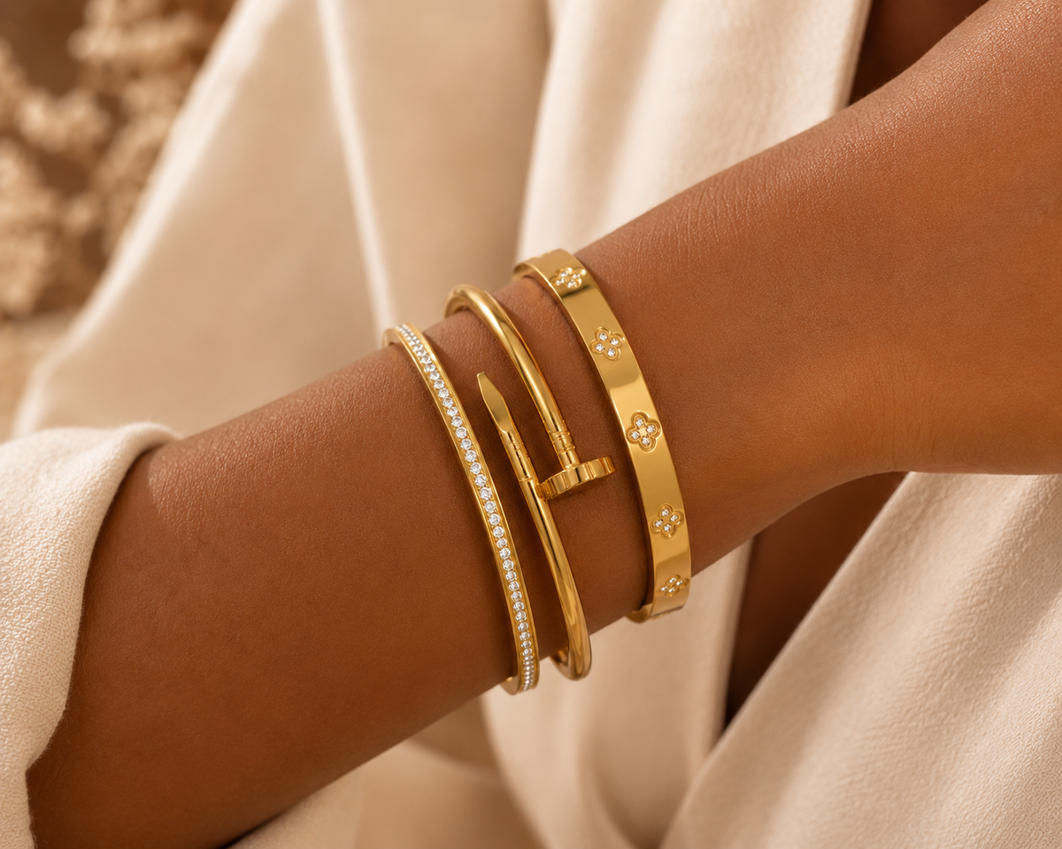 A close-up of a person's wrist wearing multiple gold bangle bracelets, including a slim gold band with small star motifs and a gold bracelet lined with clear stones, styled with a cream-colored sleeve.