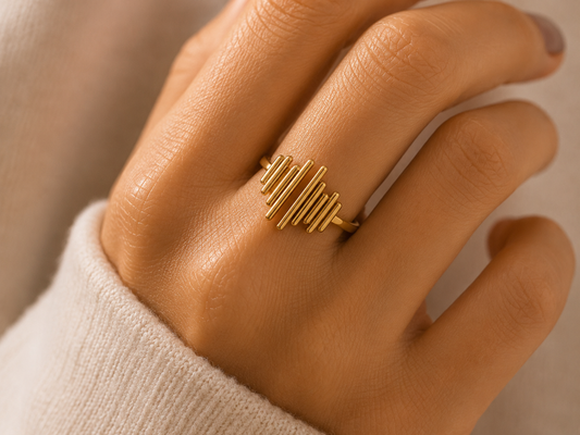 A close-up of a hand wearing a gold ring with a modern, wavy geometric design. The hand is resting on a soft, cream-colored knit sleeve against a warm, neutral background.