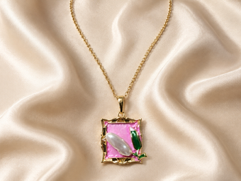 A gold pendant necklace featuring a square pink enamel background with a white mother-of-pearl bead and green enamel leaf details, displayed on a draped cream satin fabric.
