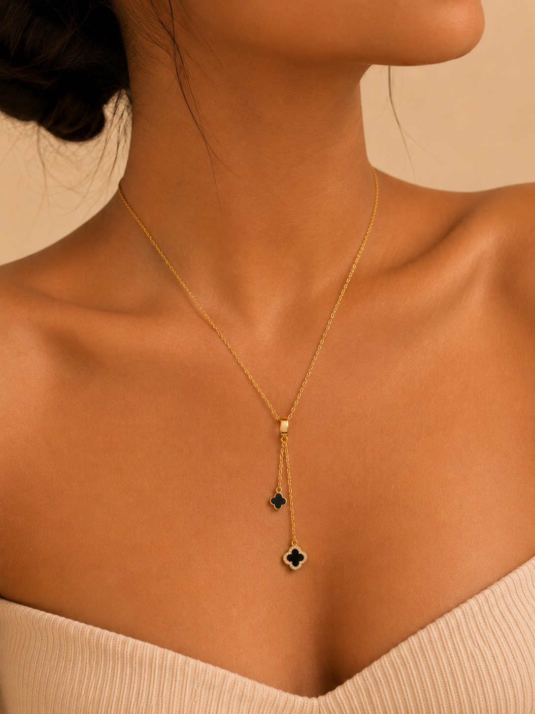 A close-up of a model wearing a delicate gold lariat necklace with two small black diamond-like drops, one of which forms a clover shape pendant.