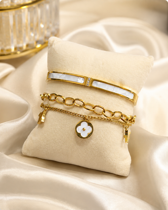 A stack of three gold-toned bracelets displayed on a cream fabric jewelry pillow. The top bracelet is a slim bangle with a white inlay, the middle is a gold chain with oval links, and the bottom is a delicate chain with a white flower charm outlined in gold.