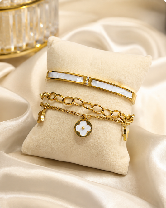 A stack of three gold-toned bracelets displayed on a cream fabric jewelry pillow. The top bracelet is a slim bangle with a white inlay, the middle is a gold chain with oval links, and the bottom is a delicate chain with a white flower charm outlined in gold.