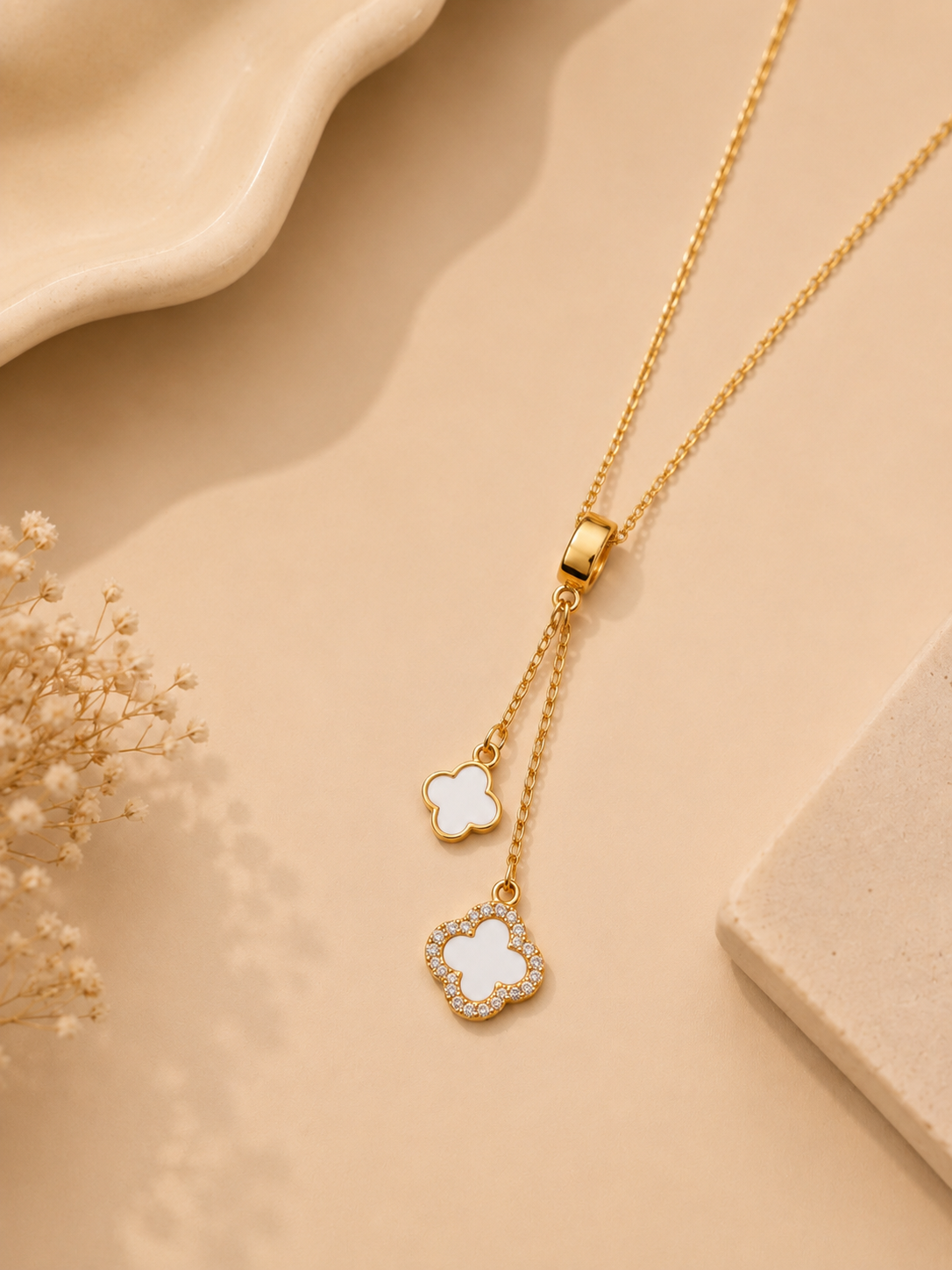 A gold-tone lariat necklace with two dangling clover-shaped pendants, one inlaid with white mother-of-pearl and the other encrusted with small clear stones, displayed on a warm beige background with dried floral accents.