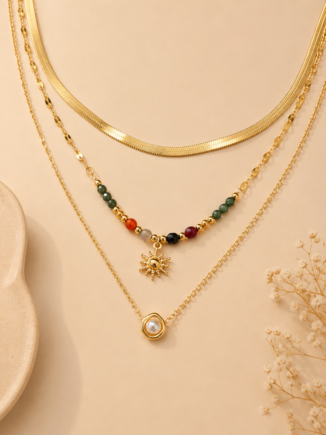 Three layered gold necklaces displayed on a beige background. The top layer features a thick gold snake chain, the middle layer has a beaded chain with a gold sun pendant, and the bottom layer has a delicate chain with a gold ring and white pearl pendant.