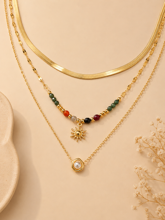 Three layered gold necklaces displayed on a beige background. The top layer features a thick gold snake chain, the middle layer has a beaded chain with a gold sun pendant, and the bottom layer has a delicate chain with a gold ring and white pearl pendant.