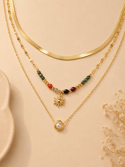 Three layered gold necklaces displayed on a beige background. The top layer features a thick gold snake chain, the middle layer has a beaded chain with a gold sun pendant, and the bottom layer has a delicate chain with a gold ring and white pearl pendant.