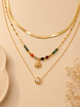 Three layered gold necklaces displayed on a beige background. The top layer features a thick gold snake chain, the middle layer has a beaded chain with a gold sun pendant, and the bottom layer has a delicate chain with a gold ring and white pearl pendant.