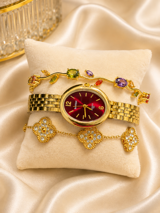 A gold-tone women's watch with a deep red dial and bezel, displayed on a cream velvet pillow over draped satin fabric. The watch is styled with two matching gold bracelets: one featuring colorful gemstone leaf clusters and another with three pavé-set clover motifs.