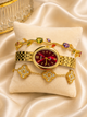 A gold-tone women's watch with a deep red dial and bezel, displayed on a cream velvet pillow over draped satin fabric. The watch is styled with two matching gold bracelets: one featuring colorful gemstone leaf clusters and another with three pavé-set clover motifs.
