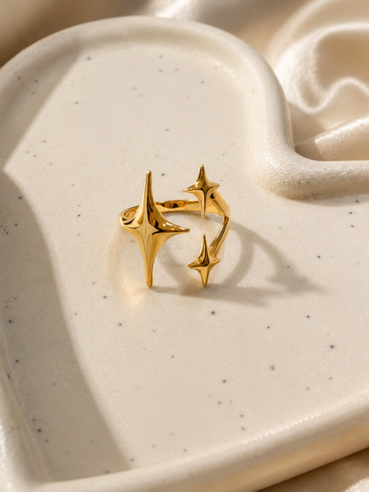 A gold-toned open ring with three starburst and crescent-like sculptural elements, displayed on a speckled cream ceramic dish with soft satin fabric in the background.