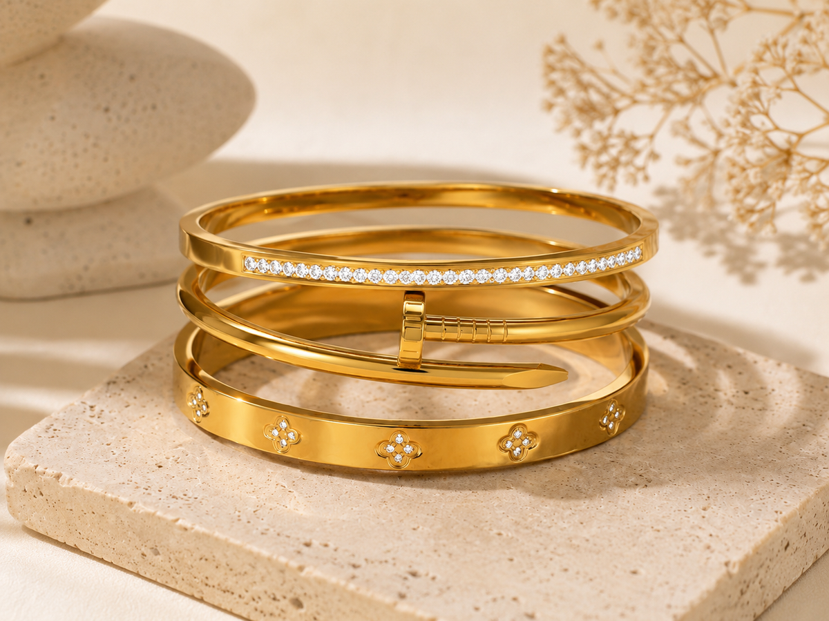 Three gold bangle bracelets stacked together on a beige stone slab, with one bracelet featuring a row of clear stones and the others accented with small diamond-like motifs. The background is softly blurred with neutral decor.