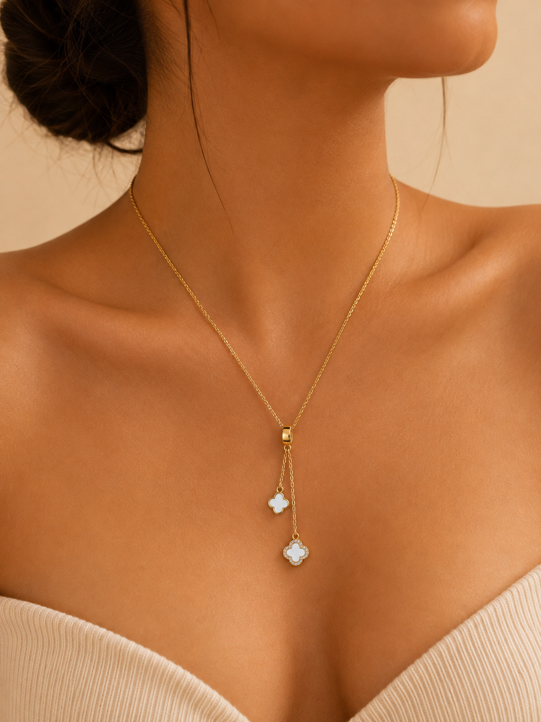 A close-up of a model wearing a delicate gold Y-necklace with two dangling charms, one small white flower and one larger white clover-like charm, set against a warm beige background.