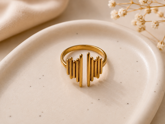 A gold-toned open ring with vertical bar elements, displayed on a round beige ceramic dish with dried flowers in the background.