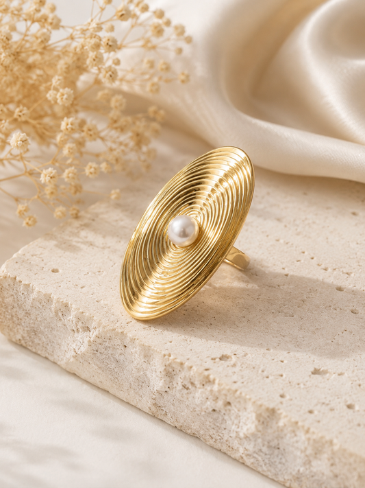 A gold-toned wide band ring with concentric circular ridges and a central white pearl, displayed on a textured stone slab with dried flowers and cream satin fabric in the background.