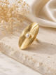 A gold-toned wide band ring with concentric circular ridges and a central white pearl, displayed on a textured stone slab with dried flowers and cream satin fabric in the background.