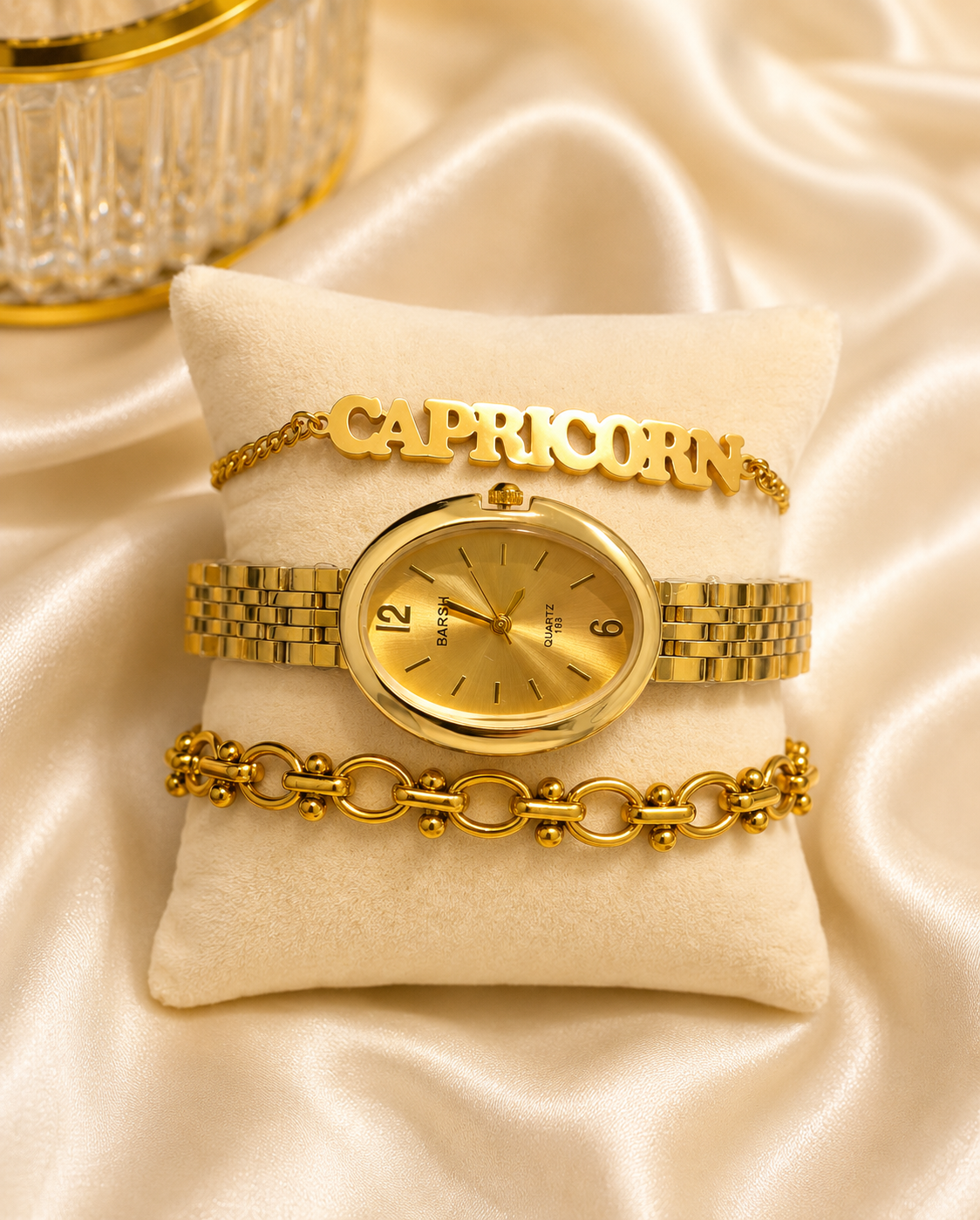 A gold-tone Capricorn bracelet watch with a matching gold-tone link bracelet and a gold-tone name bracelet spelling 'CAPRICORN', displayed on a cream velvet pillow over a champagne satin background.
