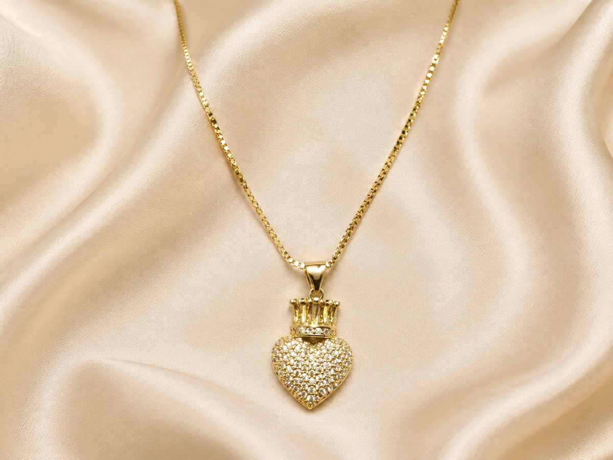 A gold heart-shaped pendant necklace covered in small clear stones, topped with a small gold crown, displayed on draped champagne satin fabric.