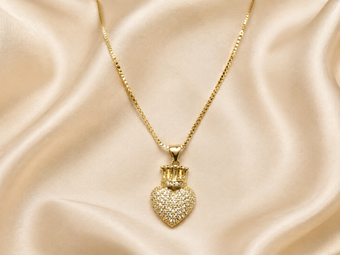 A gold heart-shaped pendant necklace covered in small clear stones, topped with a small gold crown, displayed on draped champagne satin fabric.