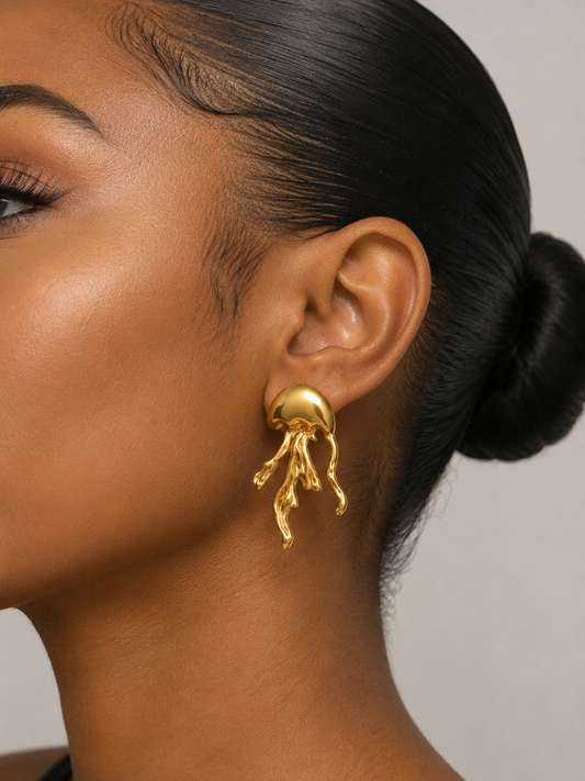 A close-up side profile of a woman wearing a gold jellyfish-shaped dangle earring. The earring features a smooth, rounded top with delicate, branching tentacles hanging below, shown against a neutral light gray background.