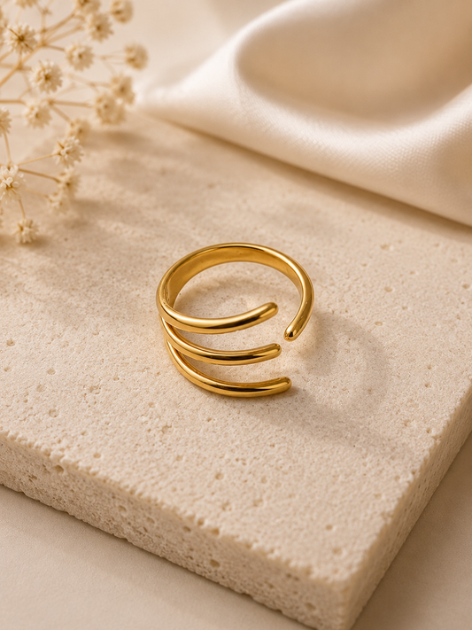 A gold-toned open bangle ring with three parallel curved bands, displayed on a textured beige stone slab with dried flowers and cream satin fabric in the background.