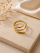 A gold-toned open bangle ring with three parallel curved bands, displayed on a textured beige stone slab with dried flowers and cream satin fabric in the background.