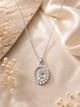 A silver oval pendant necklace with a rose motif and a border of small clear stones, displayed on a beige satin fabric with dried flowers and a textured stone prop.
