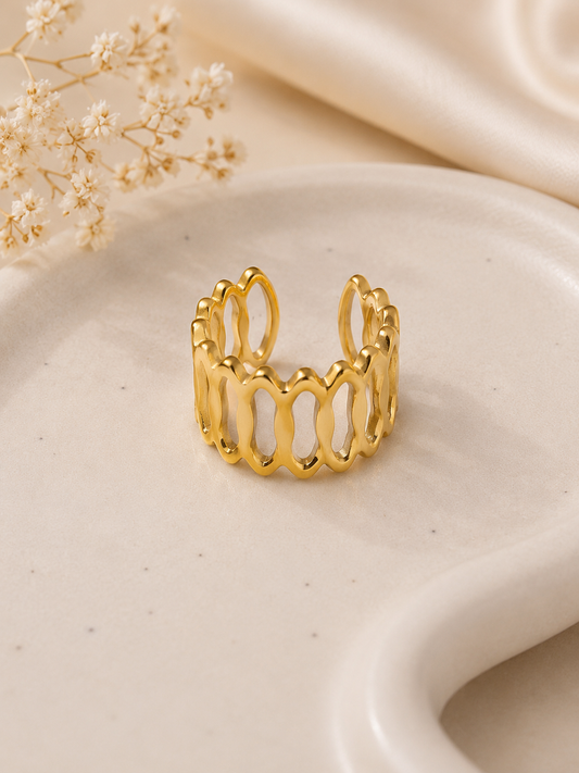 A gold open-link ring with a wavy, organic design, displayed on a beige ceramic dish with dried baby's breath flowers and a cream satin fabric in the background.