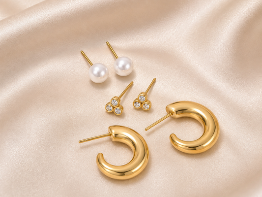 A set of gold and pearl stud earrings arranged on draped champagne satin fabric. The set includes a pair of classic white pearl studs, a pair of small gold tri-oval stud earrings with clear stones, and a pair of chunky open C-shaped gold hoop earrings.