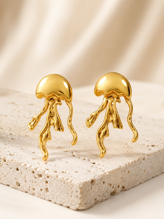 A pair of gold jellyfish stud earrings displayed on a textured beige stone slab against a soft beige fabric backdrop.