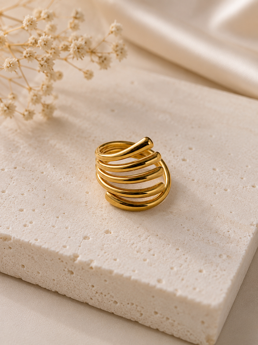 A gold-plated multi-band ring with a twisted, overlapping design, displayed on a textured beige stone block with dried flowers in the background.