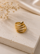 A gold-plated multi-band ring with a twisted, overlapping design, displayed on a textured beige stone block with dried flowers in the background.