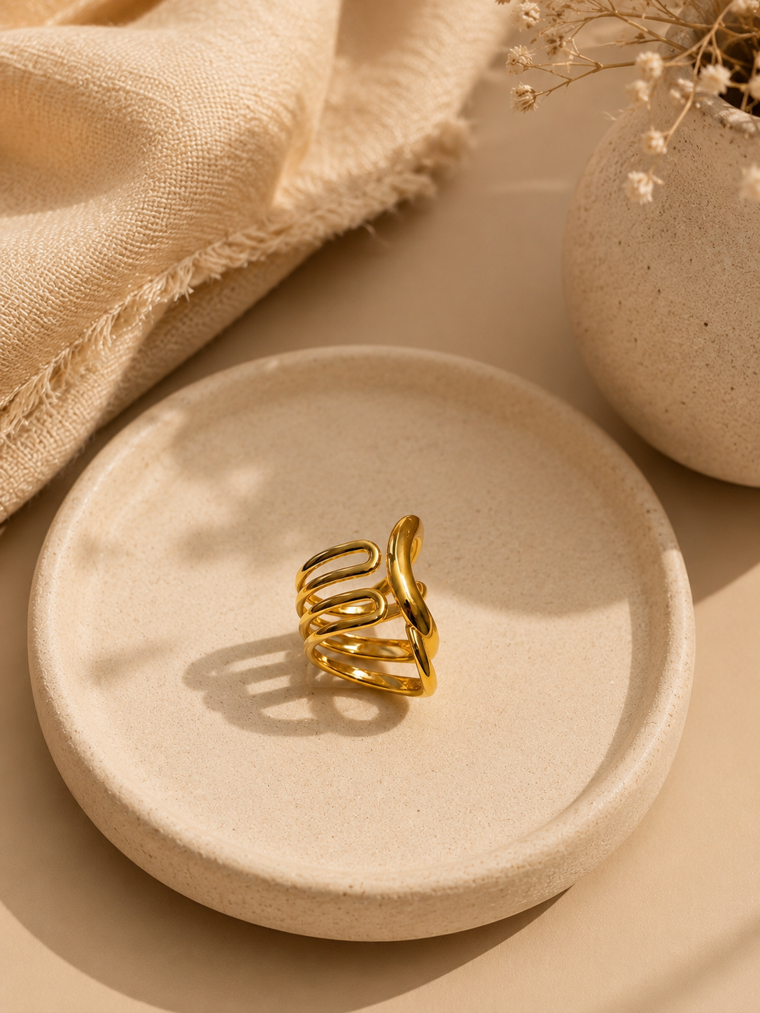 A gold-toned open spiral hair claw clip displayed on a round beige ceramic dish, styled with a textured beige fabric and dried flowers in a warm, minimalist setting.