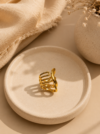 A gold-toned open spiral hair claw clip displayed on a round beige ceramic dish, styled with a textured beige fabric and dried flowers in a warm, minimalist setting.